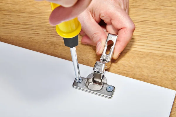 The assembly worker tightens the screw in the furniture door hinges into the wooden table made of particle board, with his other hand he holds the mechanism, ready-to-assemble furniture.