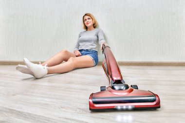 Yorgun temizlikçi kadın katta oturan ve üzerinde modern kırmızı dik elektrik süpürgesi ile led ışıkları ile toz temizleme.