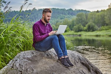 Bir genç adam doğada bir dizüstü bilgisayar kullanır.