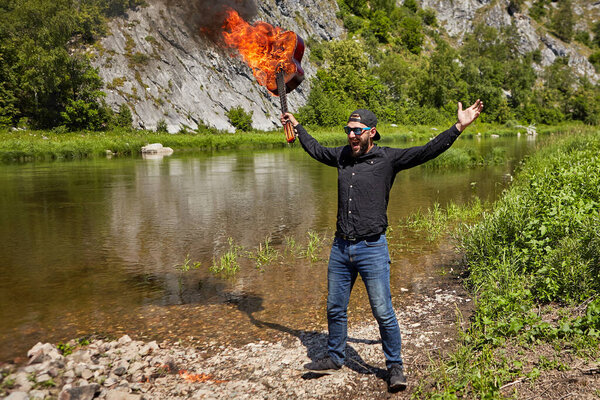 Crazy guitarist stands near river with burning guitar in his hand. A young bearded man in sunglasses and a cap, worn backwards, screams and raises burnt musical instrument. Mad musician on nature.