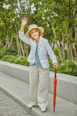 Yaklaşık 60 yaşında, beyaz bir bayan seyahat ediyor. O, yolda turuncu şemsiyeyle şapka ve günlük giysiler içinde duruyor ve gülümsüyor. Yaşlı kadın aktif ve arkadaş canlısıdır..
