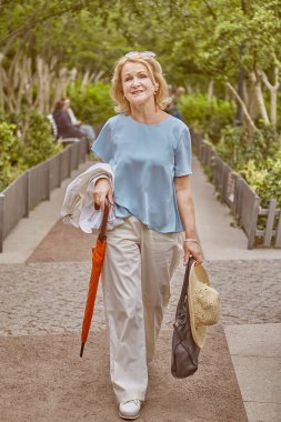 Yaklaşık 62 yaşında yaşlı, beyaz, çekici bir kadın halka açık bir parkta yürüyor. O, turuncu şemsiyeli şık giysili aktif ve neşeli bir bayan..