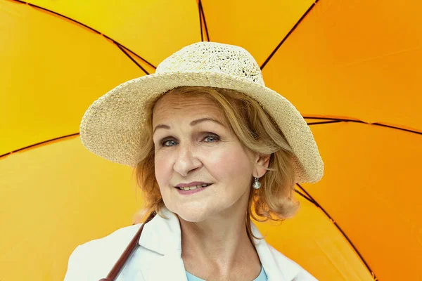 Portrait of white aged woman about 60 years old. Elderly lady is walking outdoors with orange umbrella. She has a hat on her head. She looks friendly and charming.