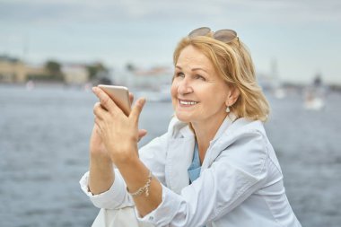 Yaşlı beyaz, güzel, gülümseyen bir bayan, 60 yaşında, elinde akıllı telefonuyla şehir merkezinde yürürken nehir kenarında duruyor. O aktif ve neşeli yaşlı bir kadın, günlük giysiler içinde..