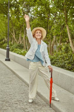 Yaklaşık 60 yaşında, aktif, beyaz, güzel bir kadın gündelik ve zarif bir elbise ve şapka ve şemsiyeyle seyahat ediyor. Yaşlı bayan halka açık bir parkta turisttir..