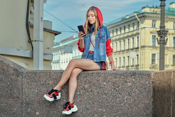 Young Woman Denim Shorts Red Hood Reads Messages Smartphone Screen — Stock Photo, Image