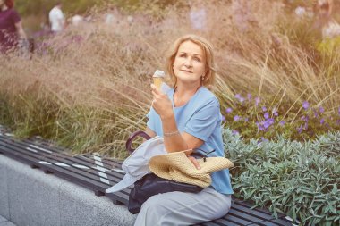 Güzel, aktif, yetişkin bir beyaz kadın, yaklaşık 62 yaşında. Coronavirus covid-19 salgını sırasında sosyal mesafeyi belirlemek için bankta oturuyor ve halka açık bir parkta dondurma yiyor..