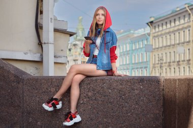 25 yaşlarında, sarışın, şortlu, kapüşonlu ve plarform ayakkabılı, kulaklıklı, Saint-Petersburg 'un ortasında akıllı telefonlu güzel bir beyaz kadın..