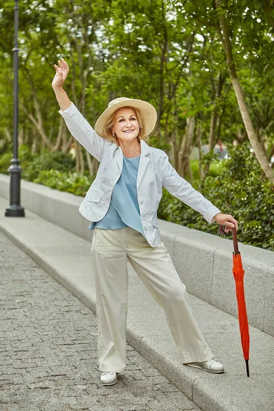 60 yaşlarında, aktif, beyaz, güzel bir bayan gündüz vakti halka açık bir parkta seyahat ediyor. Şapkası ve şemsiyesi olan sıradan ve şık giyimli oldukça olgun bir iş kadını..