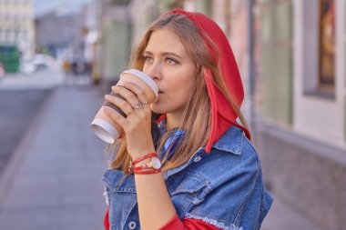 25 yaşlarında, uzun sarı saçlı, kırmızı kapüşonlu ve kot ceketli çekici beyaz kadın şehir merkezinde yürürken kağıt bardaktan kahve içiyor..