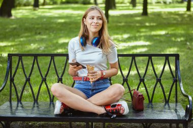 Beautiful caucasian young woman about 25 years old with blond hair and earphones is sitting on the bench in public park with mobile phone in hand and eating ice cream. clipart
