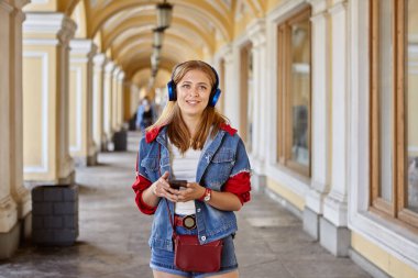 Genç bayan kemerli ticaret galerisinde telefonla konuşmak için kablosuz kulaklık kullanıyor. Güzel, beyaz, beyaz, sarı saçlı kadın yürüyor ve gülümsüyor..