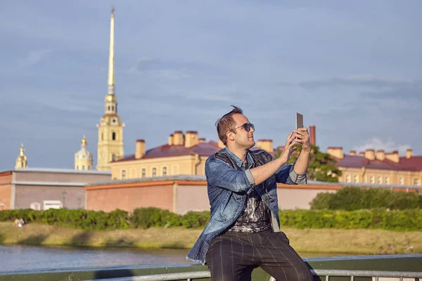 Turist, Rusya 'daki tarihi Saint-Petersburg merkezinin arka planında selfie çekiyor. Genç beyaz adam fotoğraf çekmek için akıllı telefon kullanıyor..