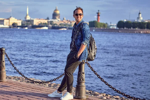 Genç adam Avrupa 'da seyahat ediyor. Güneş gözlüklü 27 yaşındaki gülümseyen beyaz erkek turist Avrupa 'da nehir kenarında oturuyor.