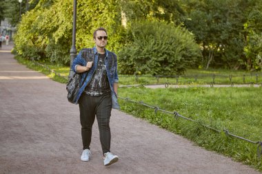 Genç adam halka açık parkta yolda yürüyor. Güneş gözlüklü beyaz erkek turist ve dışarıda sırt çantasıyla geziyor..