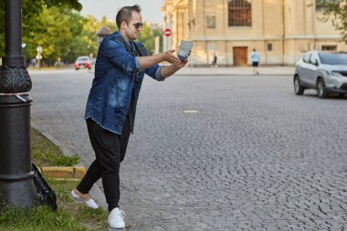Genç adam şehir caddesinde ayakkabısının kumunu sallıyor. Güneş gözlüklü beyaz erkek dışarıda yürüyor..