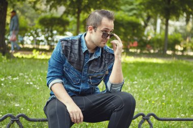 Güneş gözlüklü genç beyaz adam gündüz vakti halka açık parkta oturuyor. Kafkas erkek dışarıda yürüyor..