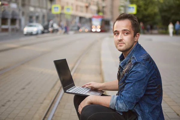 Young pensive man with notebook in hands is sitting near railroad track on the background of a riding tram.