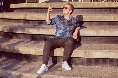 Gülümseyen adam 27 yaşında beton merdivende oturuyor ve şehirde yürürken selfie çekiyor. Genç erkek, akıllı telefon kullanarak kendi fotoğraflarını çekiyor..