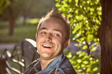 27 yaşındaki adam parktaki bankta otururken gülüyor. Yüzü batan güneşin ışınlarıyla aydınlanan gülümseyen genç bir erkek portresi..