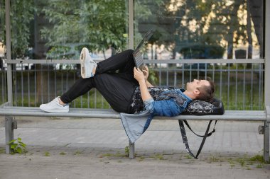 Adam otobüs durağındaki bankta dizlerinin üstünde dizüstü bilgisayarla yatıyor. Toplu taşıma istasyonunda defter taşıyan beyaz bir erkek..