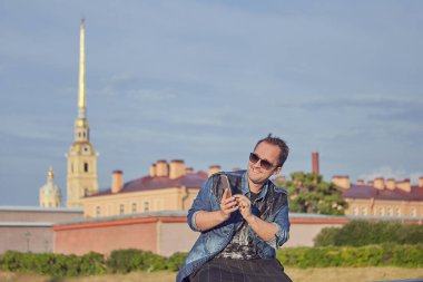 Güneş gözlüklü gülümseyen genç adam, Rusya 'nın Saint-Petersburg kentinin tarihi mimarisinin arka planında oturuyor..
