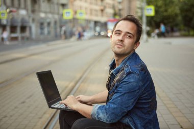 Adam dizlerinin üzerinde dizüstü bilgisayarla demiryolunun yanında oturuyor. Not defterli genç bir erkek..