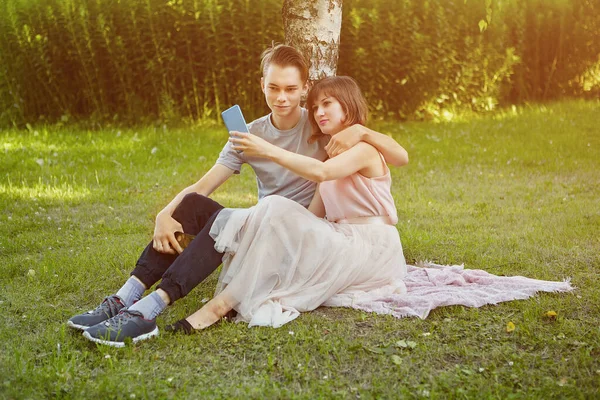 23歳の若い魅力的な男性と女性が公園の芝生の上に座っている間 スマートフォンで自撮りしています — ストック写真