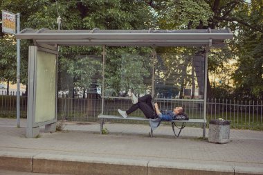 Otobüs durağındaki beyaz bir adam dizinde dizüstü bilgisayarla bankta uzandı. Genç bir erkek toplu taşıma beklerken bir dizüstü bilgisayar kullanıyor..