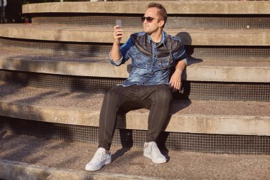 Genç bir Rus adam, güneşli bir yaz akşamında beton bir merdivenin basamaklarında otururken akıllı telefon ekranında mesajlar okur. Erkek, 27 yaşında. Açık havada sohbet etmek için telefonu kullanıyor..