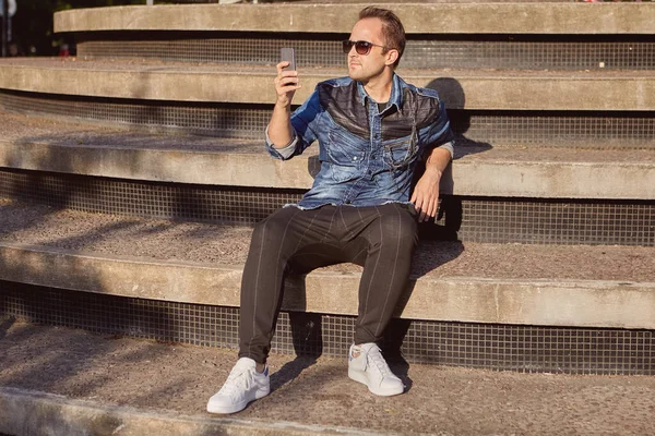 Genç bir Rus adam, güneşli bir yaz akşamında beton bir merdivenin basamaklarında otururken akıllı telefon ekranında mesajlar okur. Erkek, 27 yaşında. Açık havada sohbet etmek için telefonu kullanıyor..