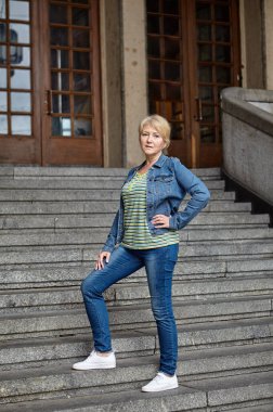 İnce, beyaz, orta yaşlı bir kadın kollarıyla antik bir binanın merdivenlerinde dikiliyor..