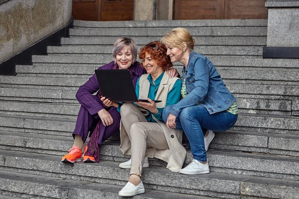 Três mulheres brancas de meia-idade sentam-se nos degraus de um edifício antigo e olham para uma tela de laptop. — Fotografia de Stock