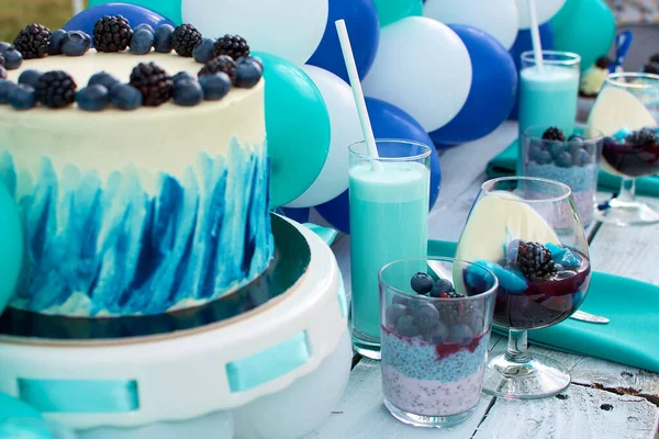 Children\'s party sweet table. Decorated with blue and white balls. Cake decorated with fresh blueberries and blackberries, milk cocktail in a tall glass, and natural milk desserts with berries