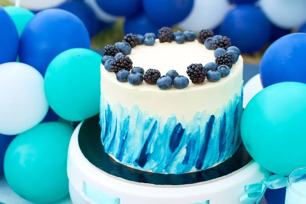 Delicious and beautiful white cake garnished with fresh blueberries and blackberries. The cake is placed on a high toad. The background is a decor of balloons. Holiday decorations in white blue colors