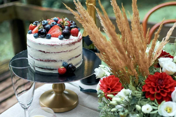 浪漫晚餐的节庆餐桌碎片 用新鲜的樱桃 草莓和蓝莓装饰的裸体蛋糕 秋天的构图 咀嚼着花朵和枯燥无味的耳朵 在后面 是一瓶起泡的酒 免版税图库照片