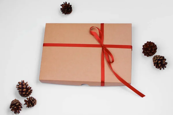 Christmas gift. Craft box with red satin ribbon on white background. Pine cones.