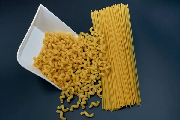 Small spring-shaped pasta is poured out of a white saucer. Spaghetti pasta on a dark background