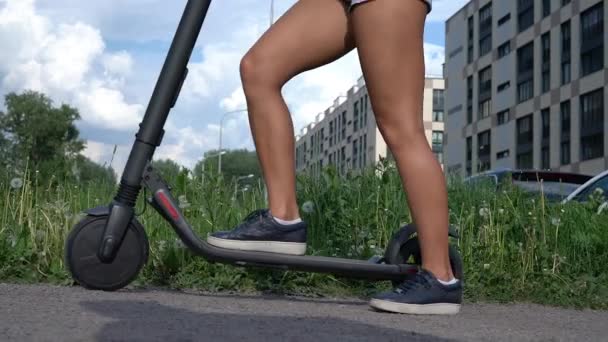 Joven mujer pasos, puestos y paseos scooter eléctrico en el fondo de hierba verde en la ciudad . — Vídeo de stock