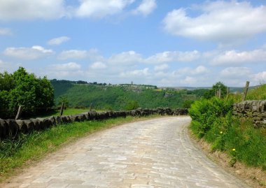 bulutlar ve heptonstall yorkshire köyü ile parlak mavi bir yaz gökyüzü ile kuru taş duvarlar ve yeşil alanlar ile çevrili uzak ormanlık vadi içine yokuş aşağı kıvrımlı bir arnavut kaldırımlı ülke şeritli