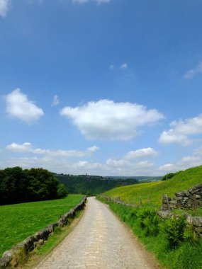 uzun arnavut kaldırımlı taş yol uzakta ormanlık vadi ve bulutlar ile parlak mavi gökyüzü ile yeşil yaz çayırları ile güzel Yorkshire dales kırsal yokuş aşağı çalışan