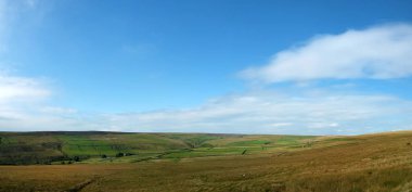 Yorkshire Dales 'in geniş panoramik manzarası. Tarlaları ve çiftlik evleri taştan duvarlarla çevrili. Üzerinde açık araziler, açık mavi gökyüzü ve bulutlar var.