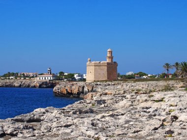 ciutadella menorca'da, derin mavi denizi ve kayalık kayalıkları ile kayalıkkayalıkların olduğu kayalıkların ve sahilin parlak güneşli manzarası, uzaktaki tarihi kale ve deniz feneri ile