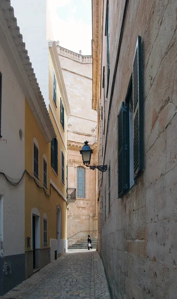 A woman walking past the end of a long quiet alley in cuitadella menorca with old historic buildings and the steps of the old cathedral in visible in the distance — Stock Photo, Image