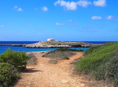 parlak mavi deniz ve yaz bulutları ile cuitadella yakınlarındaki cala santandria koyun kenarında kıyı şeridi ve eski İngiliz dönemi Torre des Castellar manzaralı bir taş yol