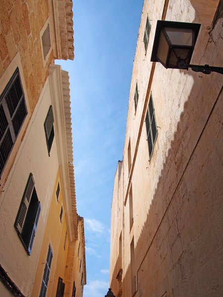 Upwards view of a typical old narrow street in ciutadella menorca with old stone buildings with shutters and street light against a bright blue sky in summer sunlight and shadow