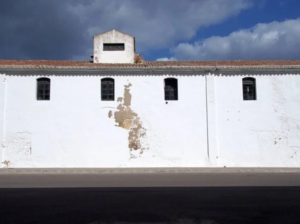 An old white painted building with high small windows and a tiled roof alongside a black roof with bright sunlit cloudy blue sky — Stock Photo, Image