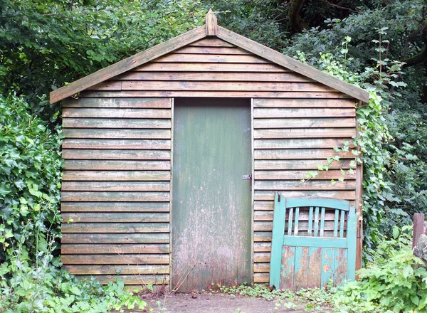 Un viejo cobertizo de madera con una puerta verde pintada y una puerta desechada apoyada en la pared rodeada de árboles y bosques con malas hierbas creciendo en el suelo — Foto de Stock