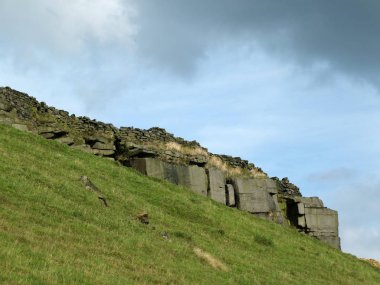 Yorkshire kırlarında dik yeşil bir yamaçta, bulutlu mavi gökyüzüne karşı tepe boyunca eski bir kuru taş duvarın kalıntılarıyla dramatik bir taş çıkıntısı