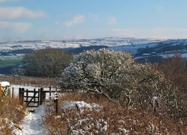 Batı Yorkshire 'da mavi gökyüzü ile parlak bir gün ışığı alan bir yol ve kapı ile kar kaplı Pennine tepeleri ve ağaçları ile panoramik bir kış sahnesi